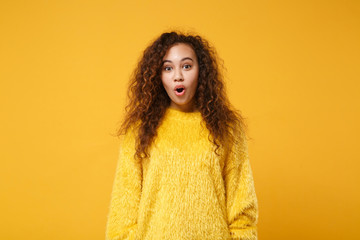 Shocked young african american girl in fur sweater posing isolated on yellow orange wall background, studio portrait. People sincere emotions lifestyle concept. Mock up copy space. Keeping mouth open.