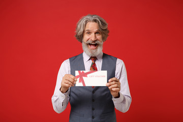 Laughing elderly gray-haired mustache bearded man in classic shirt vest and colorful tie isolated on red background in studio. People lifestyle concept. Mock up copy space. Holding gift certificate.