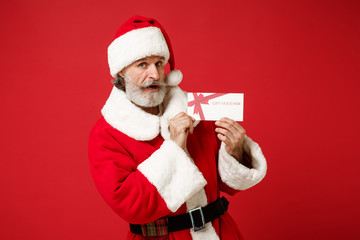 Shocked elderly gray-haired mustache bearded Santa man in Christmas hat isolated on red wall background. Happy New Year 2020 celebration holiday concept. Mock up copy space. Holding gift certificate.