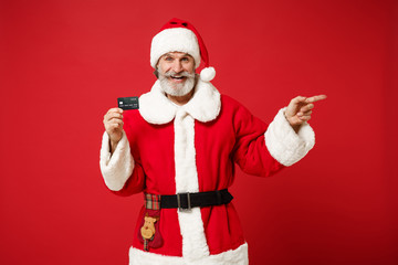 Fototapeta na wymiar Elderly gray-haired mustache bearded Santa man in Christmas hat isolated on red background. New Year 2020 celebration holiday concept. Mock up copy space. Holding credit bank card, point finger aside.