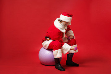Elderly gray-haired mustache bearded Santa man in Christmas hat posing isolated on red background. Happy New Year 2020 celebration holiday concept. Mock up copy space. Sit on fitball, hold dumbbells.