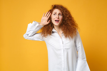 Curious young redhead woman girl in casual white shirt posing isolated on yellow orange background, studio portrait. People lifestyle concept. Mock up copy space. Try to hear you with hand near ear.