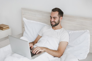 Calm young handsome bearded man lying in bed with white sheet pillow blanket in bedroom at home. Male spending time in room using pc laptop. Rest relax good mood lifestyle concept. Mock up copy space.