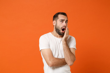 Tired young man in casual white t-shirt posing isolated on bright orange background, studio portrait. People sincere emotions lifestyle concept. Mock up copy space. Yawning, covering mouth with hand.