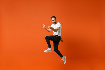 Plakat Cheerful young man in casual white t-shirt posing isolated on orange wall background. People lifestyle concept. Mock up copy space. Having fun, fooling around, jumping pointing index fingers aside.