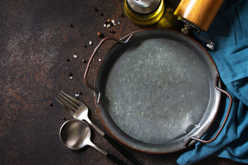 Culinary background. Round wooden metal plate, cutlery with herbs and spices around on a dark stone background from above.