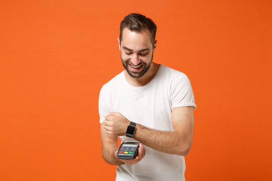 Smiling man in white t-shirt posing isolated on orange background. People lifestyle concept. Mock up copy space. Wear smart watch hold wireless bank terminal to process acquire credit card payments. - Powered by Adobe