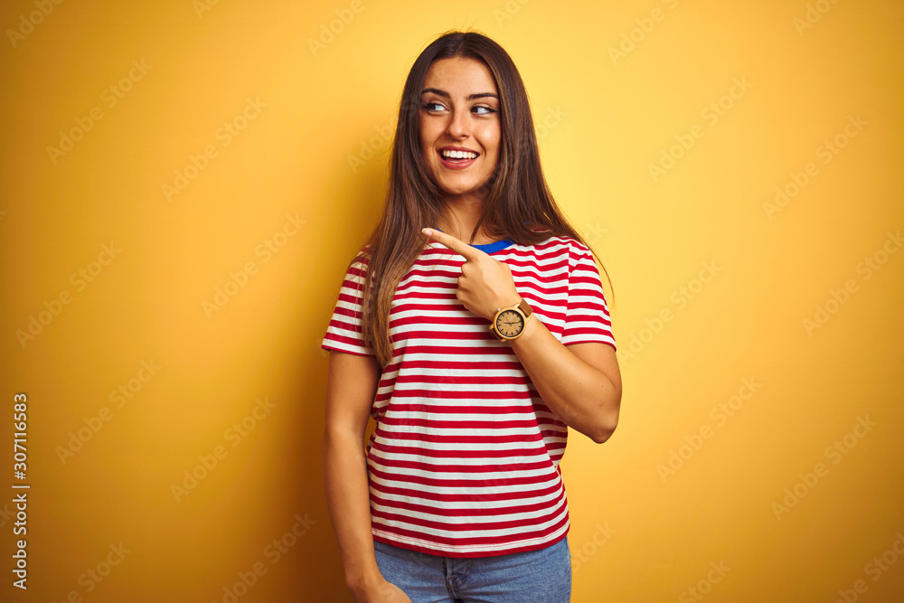 Wall mural Young beautiful woman wearing striped t-shirt standing over isolated yellow background cheerful with a smile of face pointing with hand and finger up to the side with happy and natural expression 