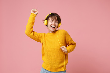 Cheerful young brunette woman girl in yellow sweater posing isolated on pastel pink wall background studio portait. People lifestyle concept. Mock up copy space. Listen music with headphones, dancing.