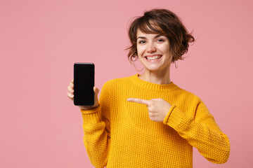 Smiling young brunette woman girl in yellow sweater posing isolated on pastel pink background. People lifestyle concept. Mock up copy space. Point index finger on mobile phone with blank empty screen.
