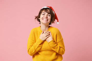 Cheerful young brunette woman Santa girl in yellow sweater, Christmas hat posing isolated on pink background. Happy New Year 2020 celebration holiday concept. Mock up copy space. Hold hands on chest.