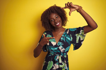 Young african afro woman wearing summer floral dress over isolated yellow background smiling making frame with hands and fingers with happy face. Creativity and photography concept.