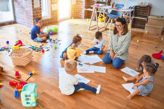 Beautiful teacher and group of toddlers sitting on the floor drawing using paper and pencil around lots of toys at kindergarten