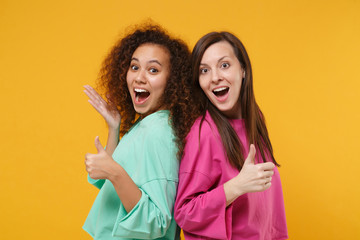 Two excited women friends european and african american girls in pink green clothes posing isolated on yellow orange wall background. People lifestyle concept. Mock up copy space. Showing thumbs up.