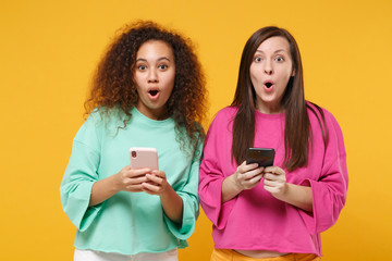 Two shocked women friends european african american girls in pink green clothes posing isolated on yellow background. People lifestyle concept. Mock up copy space. Using mobile phone, typing message.