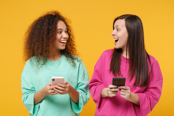 Two excited women friends european african american girls in pink green clothes posing isolated on yellow background. People lifestyle concept. Mock up copy space. Using mobile phone, typing message.