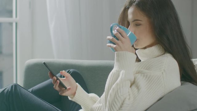 Beautiful Young Woman Using Smartphone, Drinks Tea while Sitting on the Chair. Sensual Girl Wearing Sweater Surfs Internet, Posts On Social Media, Shares Pictures while Relaxing in Cozy Apartment