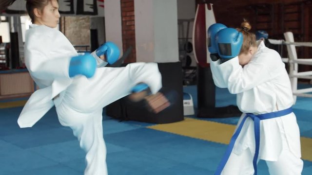 Medium shot of female Caucasian karateka wearing white kimono and blue boxing gloves fighting with her female opponent in gym