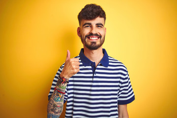 Young man with tattoo wearing striped polo standing over isolated yellow background doing happy thumbs up gesture with hand. Approving expression looking at the camera with showing success.
