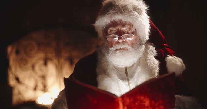 Authentic caucasian senior in santa claus clothing looking through book with red cover, sitting near his fireplace - christmas spirit, holidays and celebrations concept close up 4k