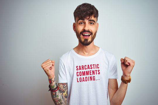 Young man with tattoo wearing fanny t-shirt standing over isolated white background celebrating surprised and amazed for success with arms raised and open eyes. Winner concept.