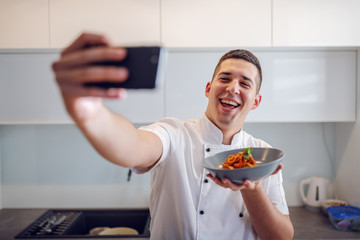 Smiling caucasian proud chef in white uniform holding plate with tagliatelle and taking selfie while standing in kitchen. - Powered by Adobe