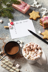 A cup of hot cocoa with marshmallows, Christmas sweets and a to-do list on a stone countertop.
