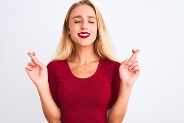 Young beautiful woman wearing red t-shirt standing over isolated white background gesturing finger crossed smiling with hope and eyes closed. Luck and superstitious concept.
