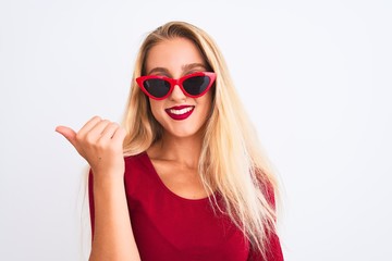 Naklejka premium Young beautiful woman wearing red t-shirt and sunglasses over isolated white background smiling with happy face looking and pointing to the side with thumb up.