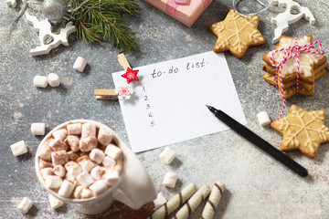 A cup of hot cocoa with marshmallows, Christmas sweets and a to-do list on a stone countertop.