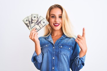 Young beautiful blond woman holding dollars standing over isolated white background surprised with an idea or question pointing finger with happy face, number one