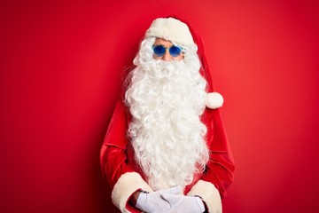 Middle age man wearing Santa Claus costume and sunglasses over isolated red background happy face smiling with crossed arms looking at the camera. Positive person.
