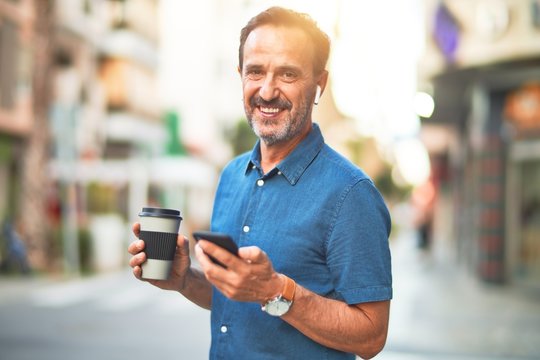 Middle age handsome businessman standing on the street using smartphone and earphones drinking coffee