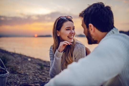 Cute smiling Caucasian brunette sitting on coast near river with her loving boyfriend and flirting. In background is sunset.