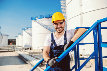Smiling handsome caucasian worker in overall and with helmet on head leaning on the railing. Oil...