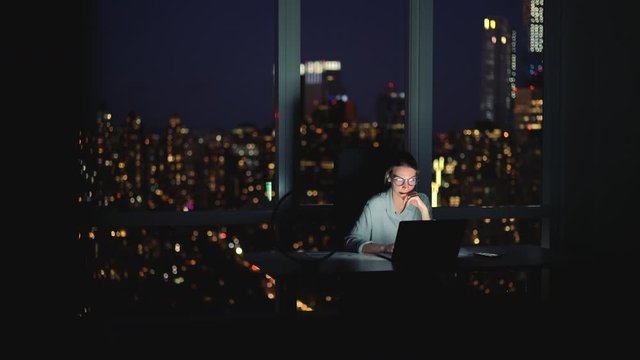 Successful woman in classic spectacles for eyes protection working at laptop device during evening extra hours in office interior with cityscape lights in skyscraper window