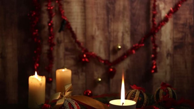 Mood of Christmas setting: one candle with big flame in foreground, red and gold baubles, decoupage balls, satin gold ribbon on tartan tablecloth on wooden background filmed from top to bottom