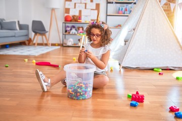 Beautiful toddler wearing glasses and unicorn diadem sitting on the floor playing with building blocks at kindergarten