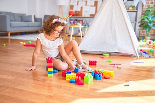 Beautiful toddler wearing glasses and unicorn diadem sitting playing with building blocks at kindergarten