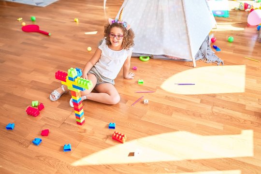 Beautiful toddler wearing glasses and unicorn diadem sitting playing with building blocks smiling at kindergarten