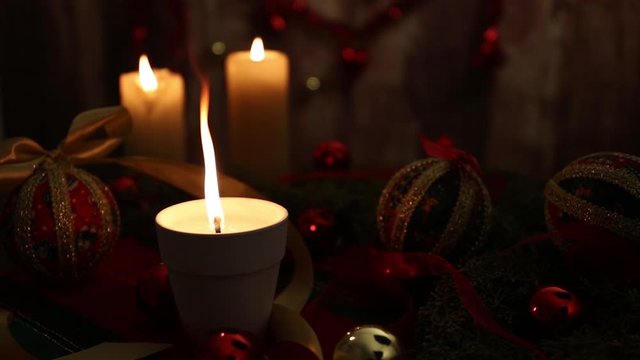 Mood of Christmas: three lit candles, one with big flame in foreground, red and gold baubles, decoupage balls, satin gold ribbon, Christmas decoration on wooden background filmed in bokeh effect