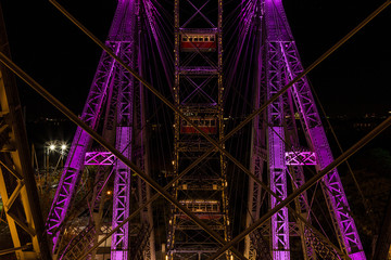 Three empty cars behind several steel wires on a giant ferris wheel covered in purple and yellow light