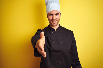 Young chef man wearing uniform and hat standing over isolated yellow background smiling friendly offering handshake as greeting and welcoming. Successful business.