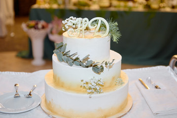 Elegant white wedding cake decorated with flowers, green leaves and golden glitter on table in restaurant, copy space