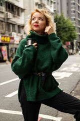 Fototapeta premium pretty young blond smiling girl in green sweater on the street hold cup in her hands outside in day time