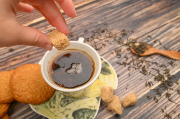 The girl's hand puts a piece of brown sugar in a mug of black tea, oatmeal cookies, tea leaves, brown sugar on a wooden background. Close up.