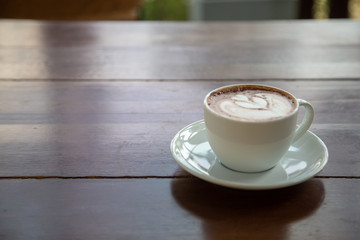 A white cup of coffee placed on a wooden table by the window in a coffee shop, Concept: Delicious breakfast drinks fragrant suitable for lifestyle, closeup cup of coffee over the top with space