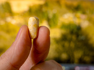 Fingers holding herbal pill against greenery defocused in the background