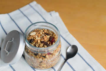 granola with peanuts, hazelnuts, oat and wheat flakes in glass jar on striped textile napkin on wooden table, close up view from angle above of horizontal still life stock photo image
