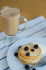 small circular fritter or pancakes with black raspberry, smetana, fork on plate and cappuccino on striped napkin, russian breakfast, closeup view from above of vertical life style stock photo image